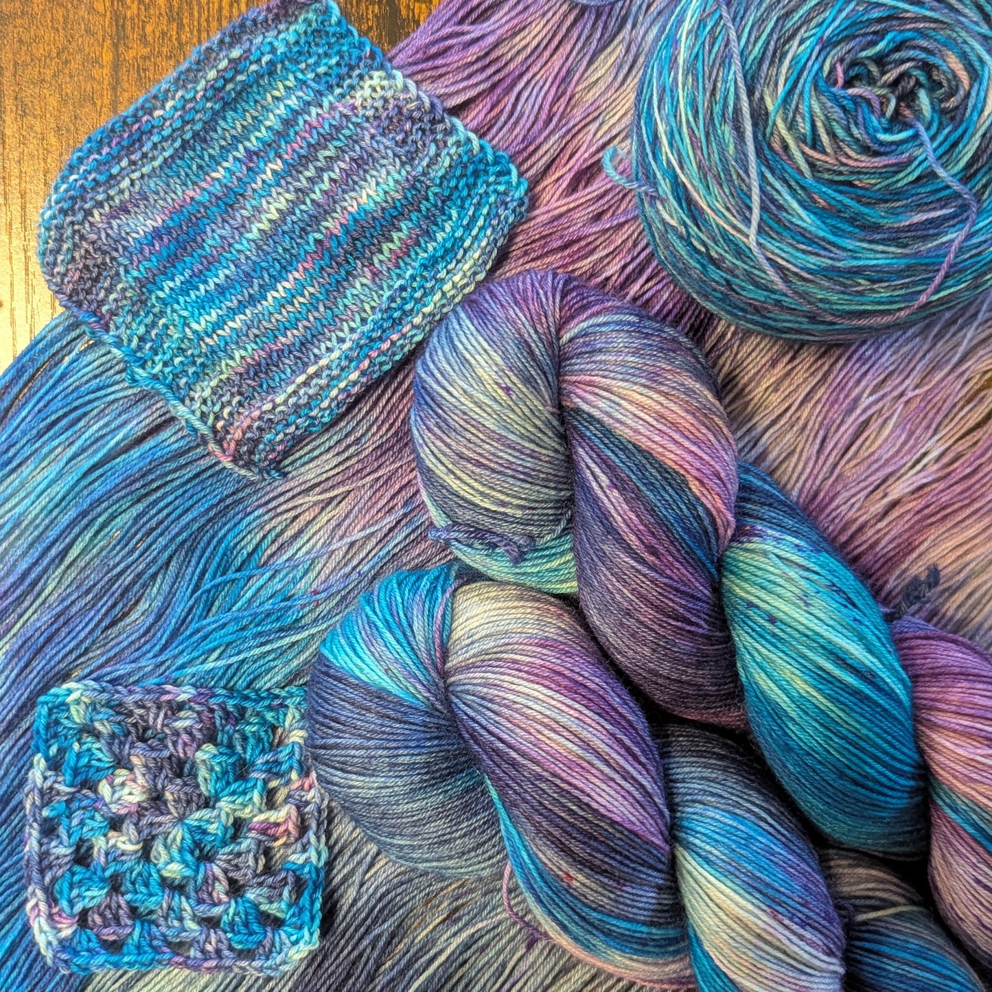 Colorful variegated yarn skeins and a knitted swatch displayed on a rustic wooden surface, showing shades of blue, purple, and teal.