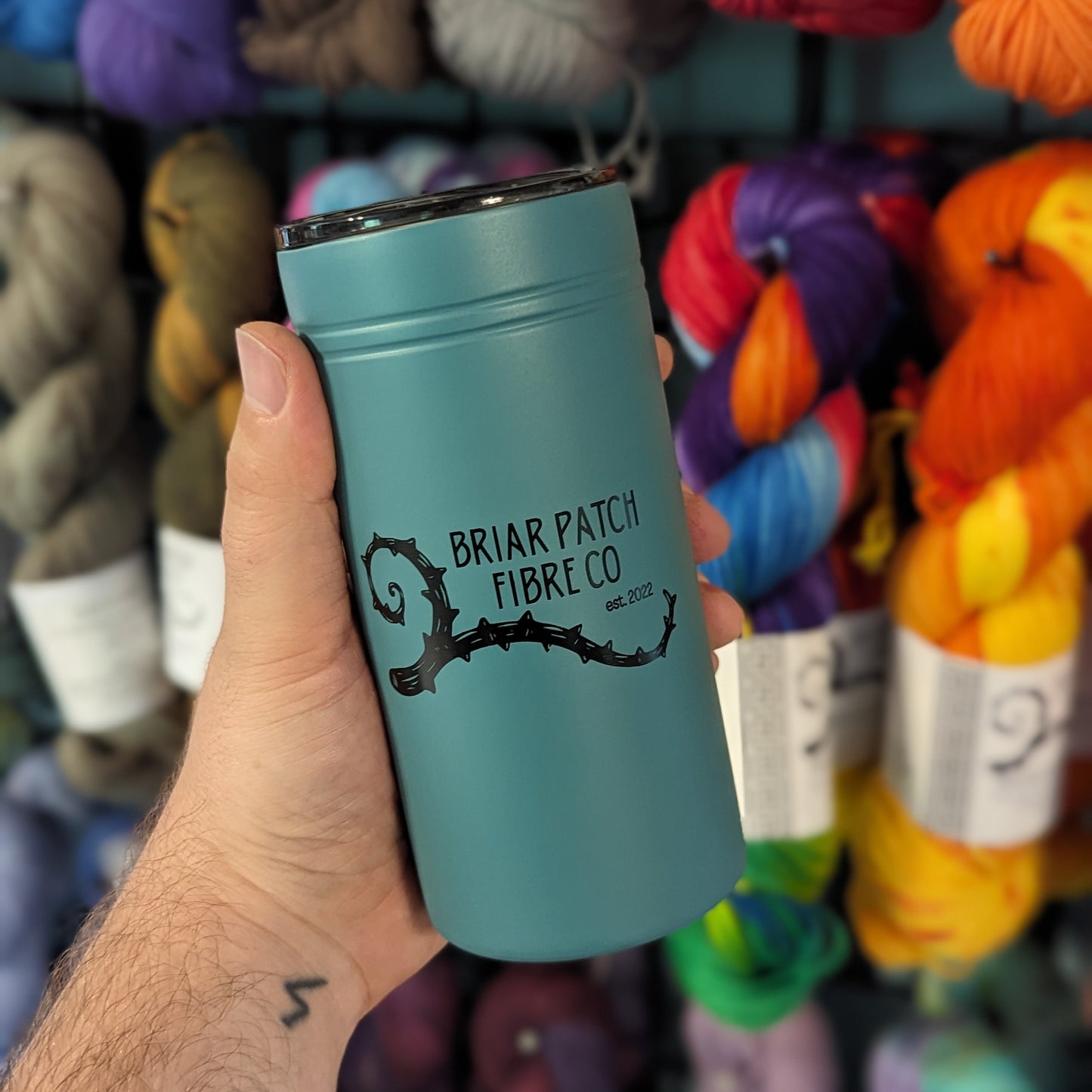 Teal tumbler with 'Briar Patch Fibre Co' logo held in front of colorful yarn balls.